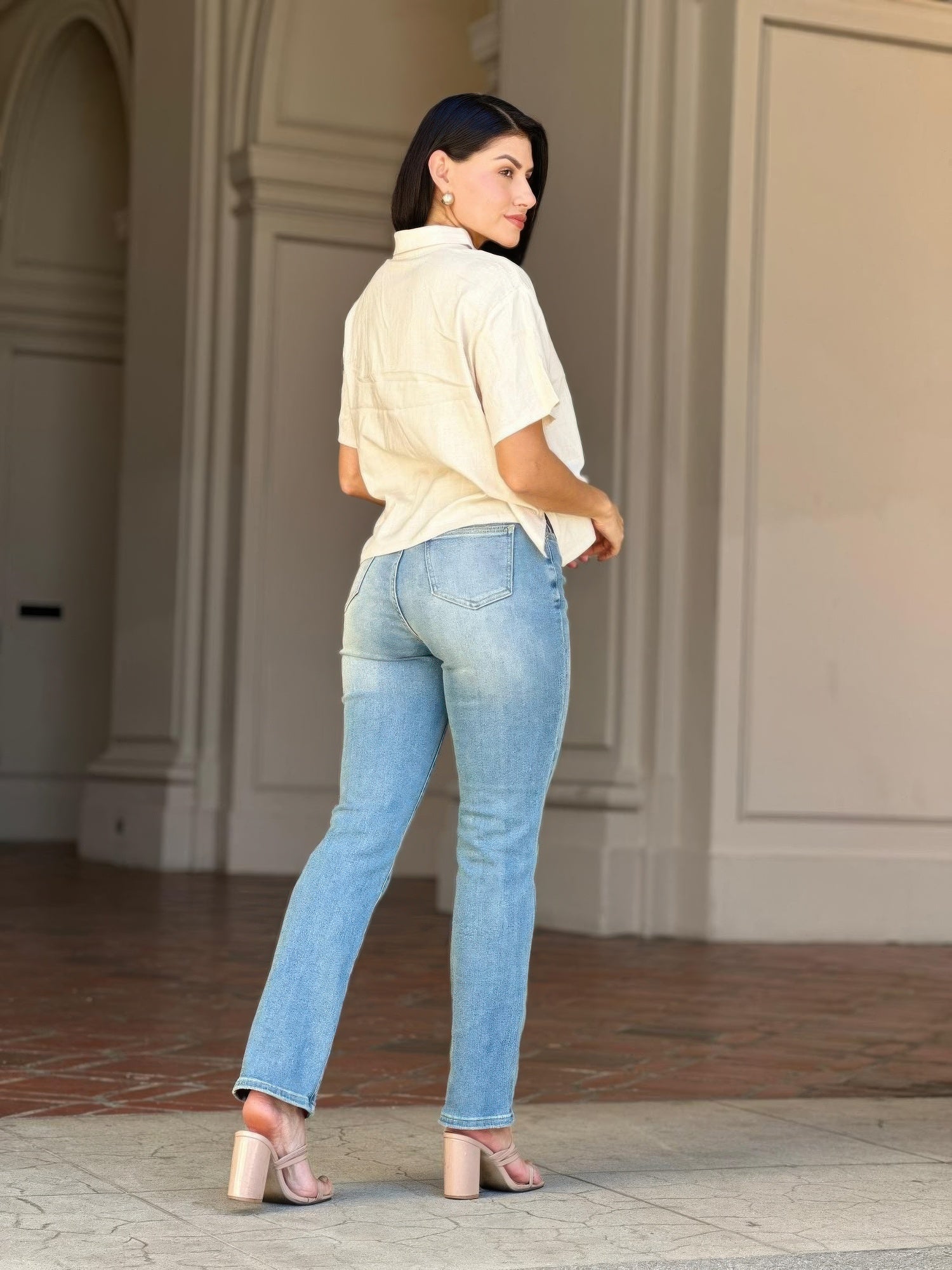 Woman in High Rise Cigarette Button Fly Straight Jeans and a cream shirt, looking over her shoulder