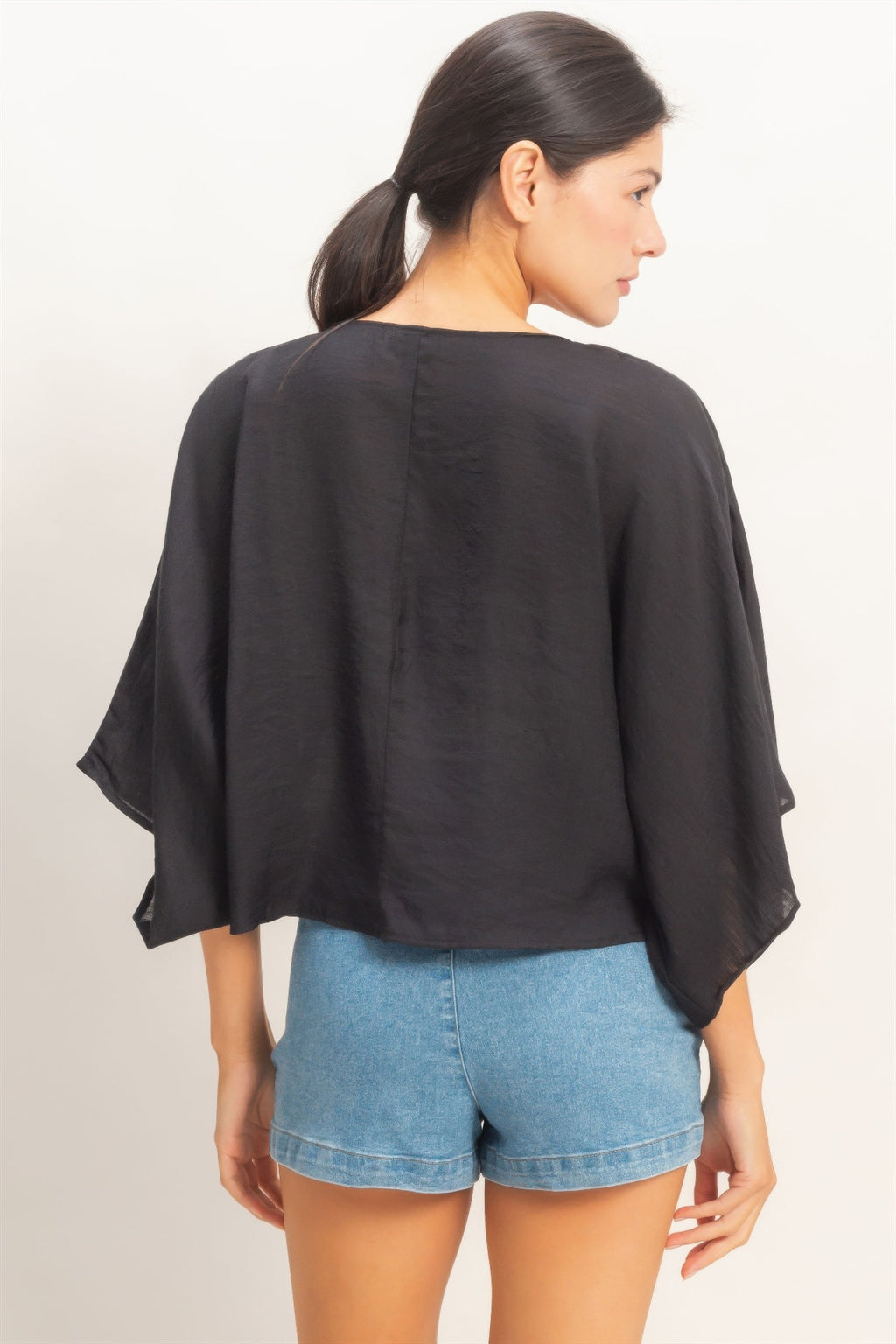 Rear view of a woman wearing an Ink Black Twist-Front Draped Shrug Top and blue denim shorts