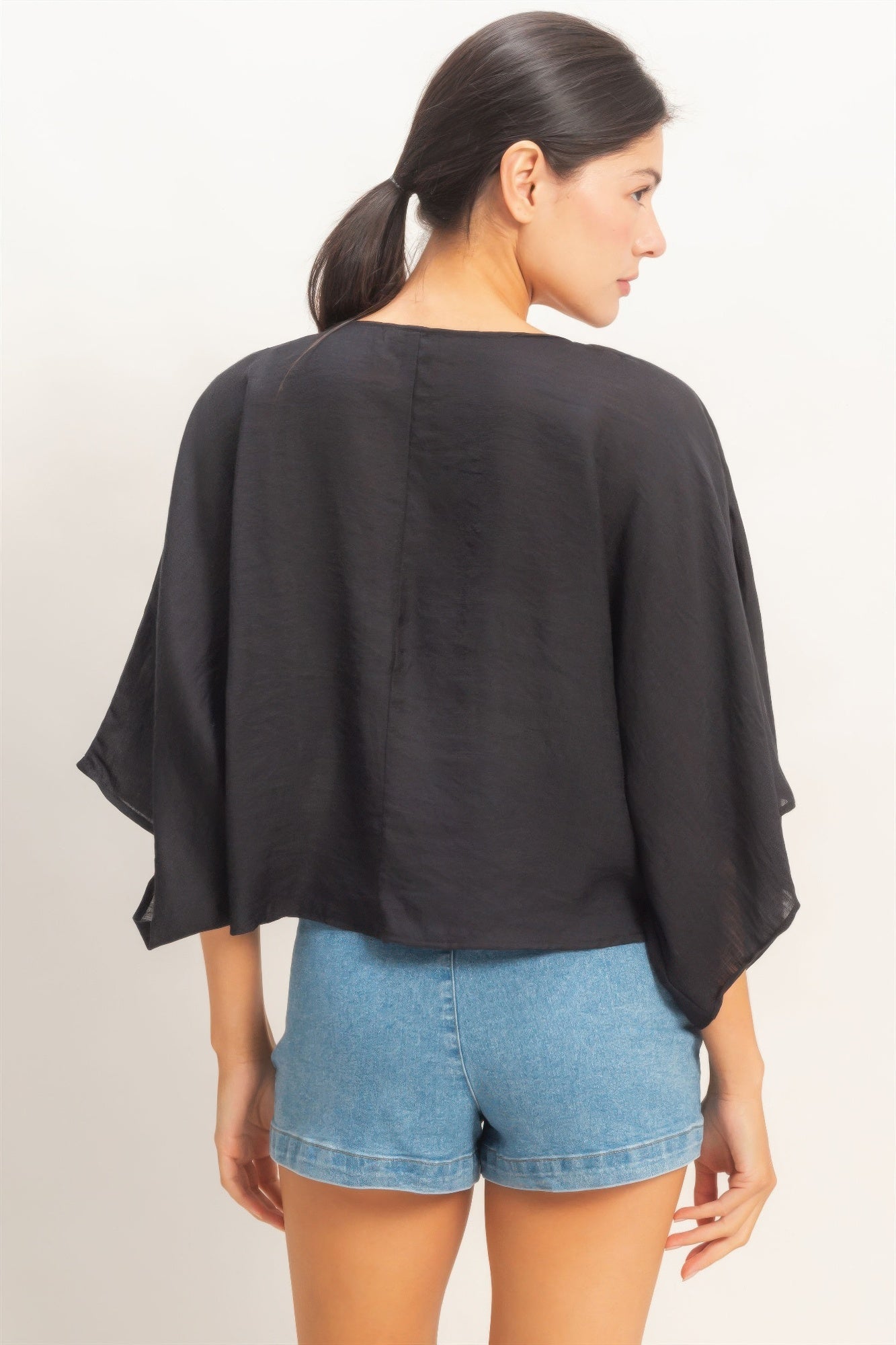 Rear view of a woman wearing an Ink Black Twist-Front Draped Shrug Top and blue denim shorts