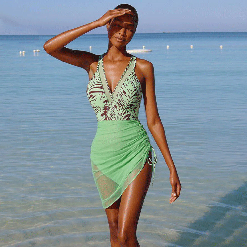 Woman in a green printed one-piece swimsuit and mesh skirt standing in shallow ocean water
