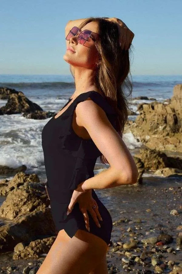 Davi & Dani Solid One Shoulder Ruffle One Piece Swimsuit in black, worn by a woman on a rocky beach